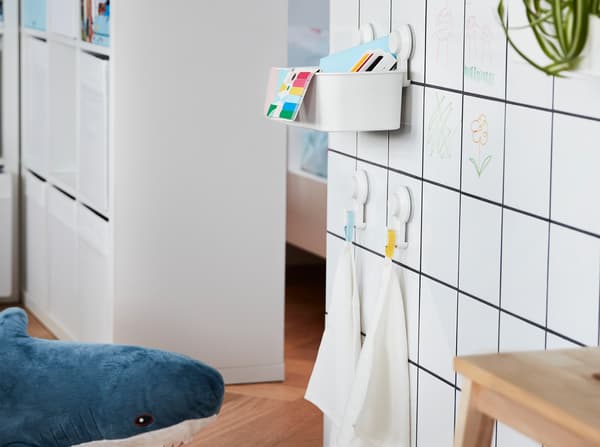 Una pared de baldosas blancas con una canasta TISKEN y ganchos con ventosas adheridas, contienen juguetes para niños y toallas pequeñas.