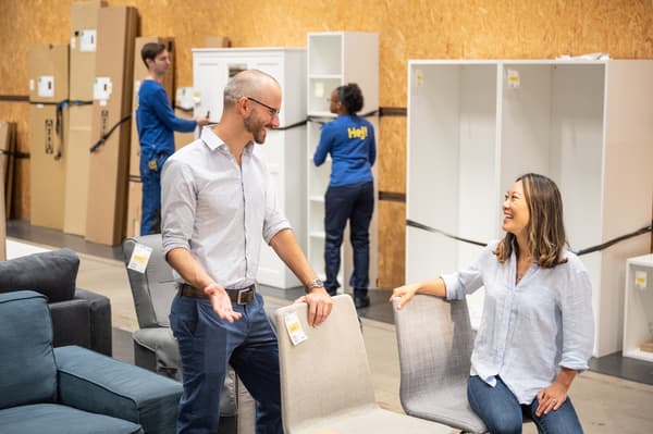 Una pareja conversando y viendo sillas en una tienda IKEA, de fondo un par de colaboradores moviendo muebles.