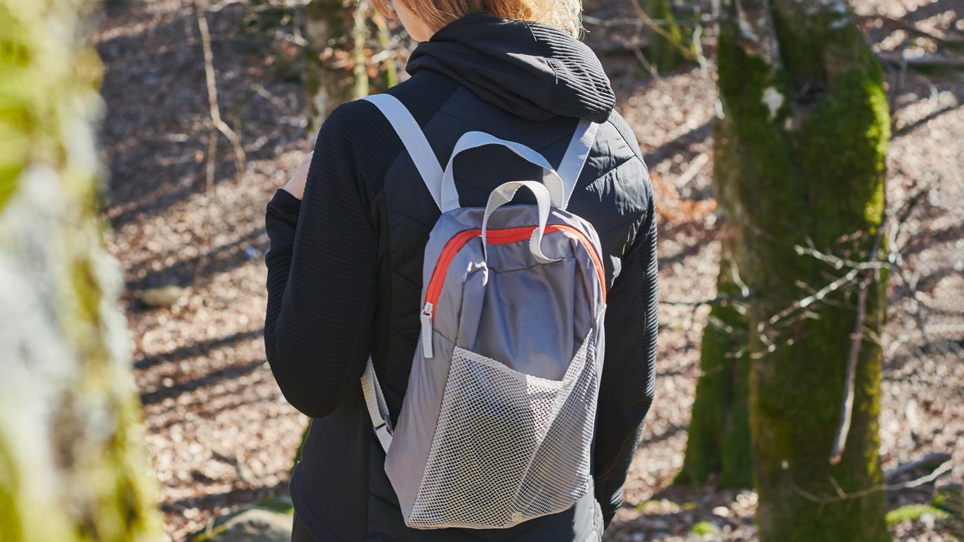 Una persona camina bajo una ladera iluminada por el sol y escasamente arbolada, con una chamarra y una mochila PIVRING de color gris claro.