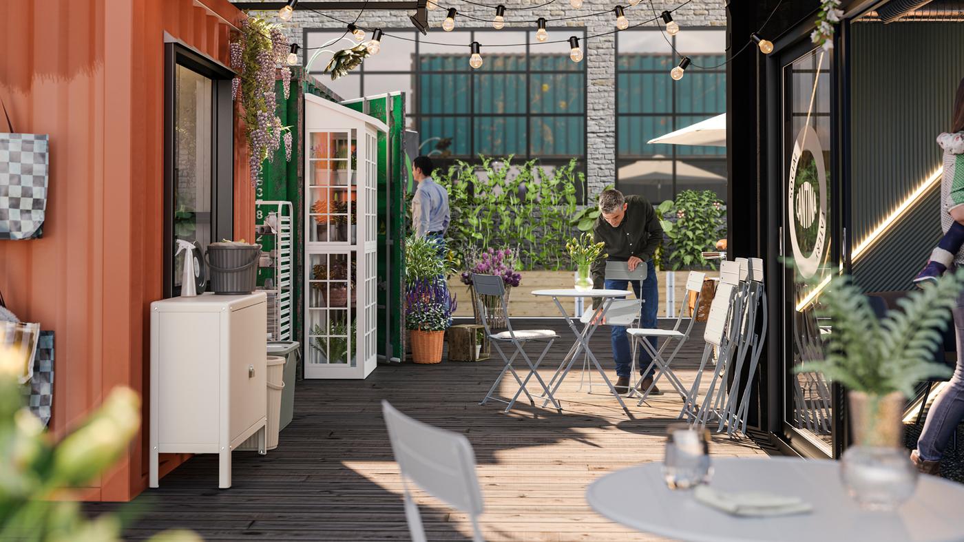 Una persona con una camicia nera sistema una sedia SUNDSÖ grigia in un'area caffè esterna decorata con numerose piante - IKEA.