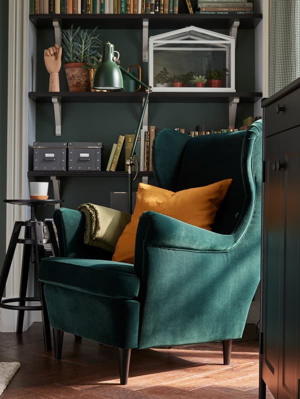 Una poltrona STRANDMON verde scuro con un cuscino ocra, una lampada da terra con il braccio ad angolo e scaffali con libri e piante - IKEA