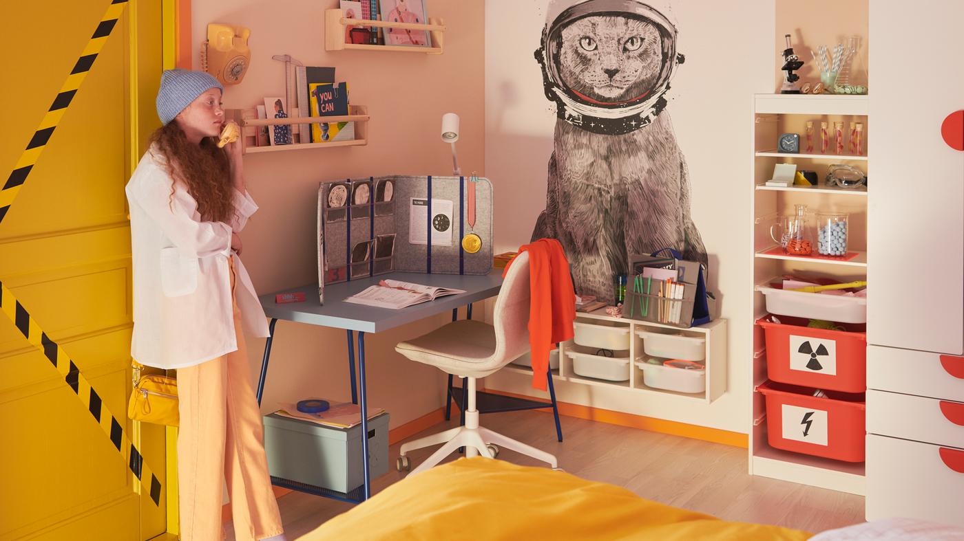 Una ragazza vicino alla porta di una stanza dai colori pastello con una scrivania LAGKAPTEN/TILLSLAG blu, una sedia da ufficio LÅNGFJÄLL, mobili e un letto - IKEA