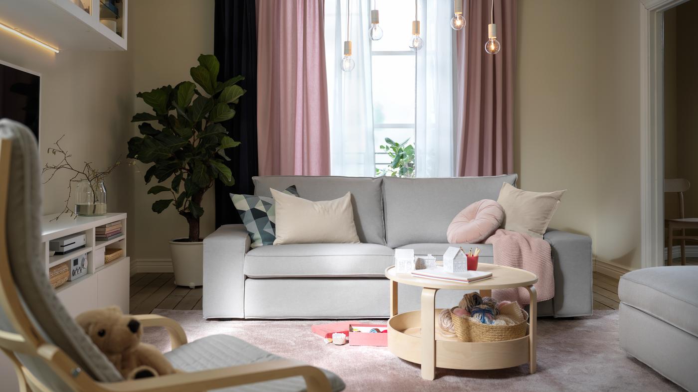 Una sala en tonos pastel con un sofá KIVIK gris claro, cortinas KALKFLY rosa claro y una mesa de centro BORGEBY.