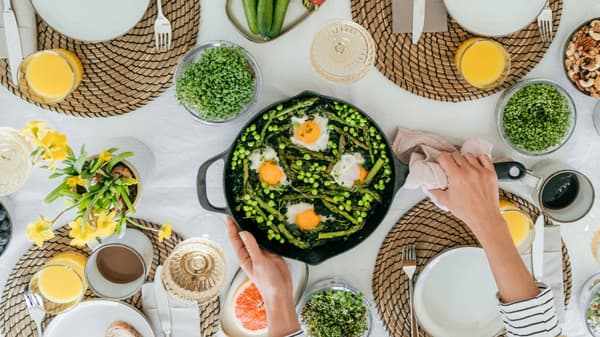 Una tavola imbandita per il brunch, con una padella contenente uova, asparagi e piselli, circondata da piatti, bicchieri di succo d’arancia e verdure fresche. Una mano sta servendo il piatto.