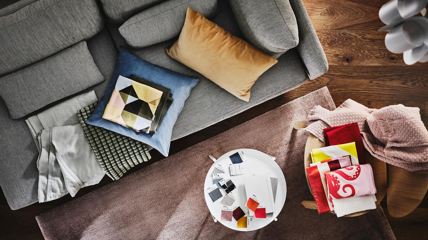Una vista dall'alto di un divano grigio con coperte e cuscini colorati. Vicino c'è un tavolino con campioni di tessuti - IKEA.