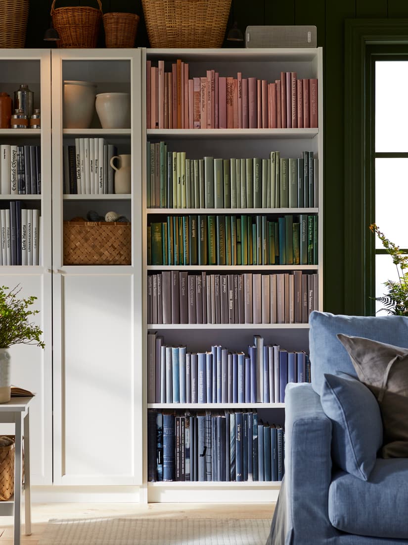 Une bibliothèque blanche BILLY remplie de livres organisés par couleur en dégradé arc-en-ciel, avec des paniers et vases décoratifs à gauche et un canapé bleu partiellement visible à droite.