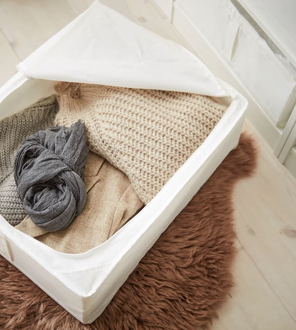 Une boîte de rangement blanche SKUBB remplie de textiles dans des tons et des fibres naturels, posée sur un tapis en fourrure brun.