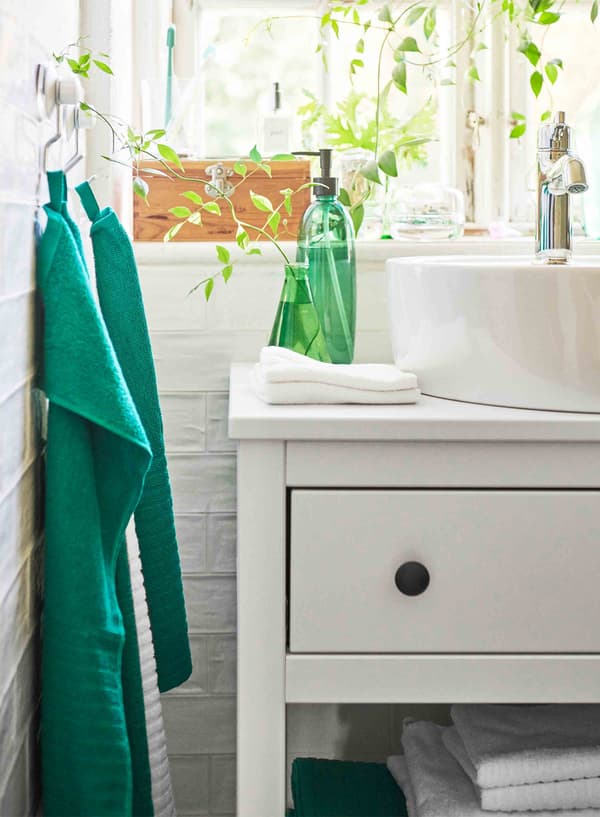 Une bouteille et un vase vert vif sont posés sur un meuble lavabo blanc, et deux serviettes vertes sont accrochées à gauche.