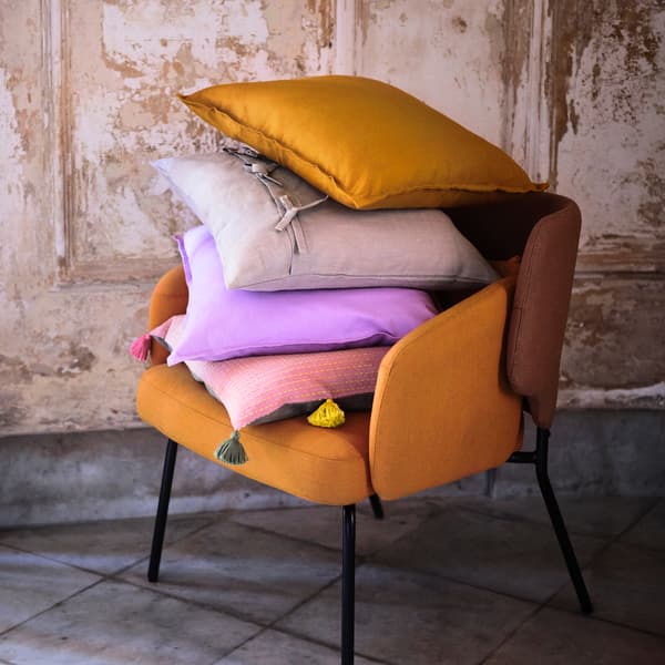 Une chaise couleur citrouille, avec différents coussins en mauve, rose et gris, posée contre un mur beige rustique.