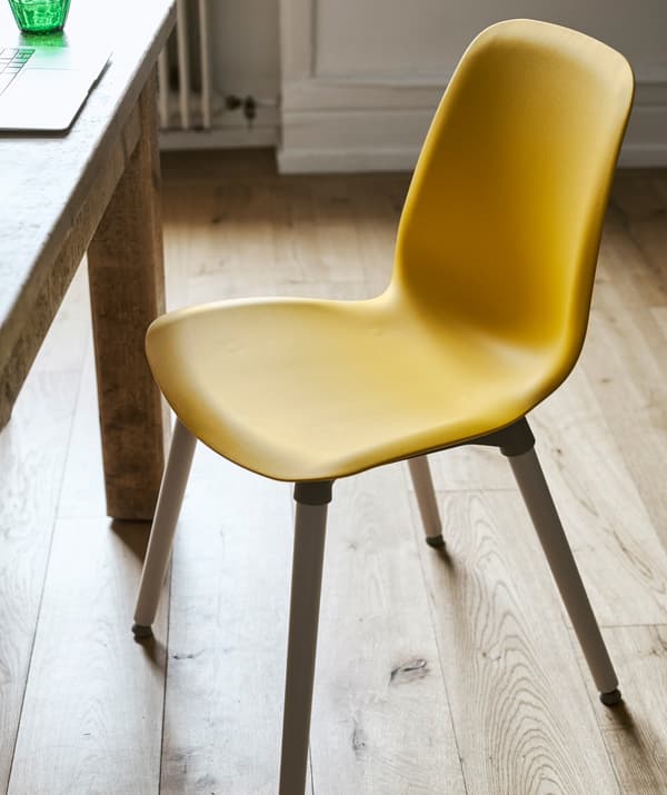 Une chaise de salle à manger avec assise en plastique jaune et pieds en bois, tirée de sous une table en bois, dans une pièce au plancher en bois.