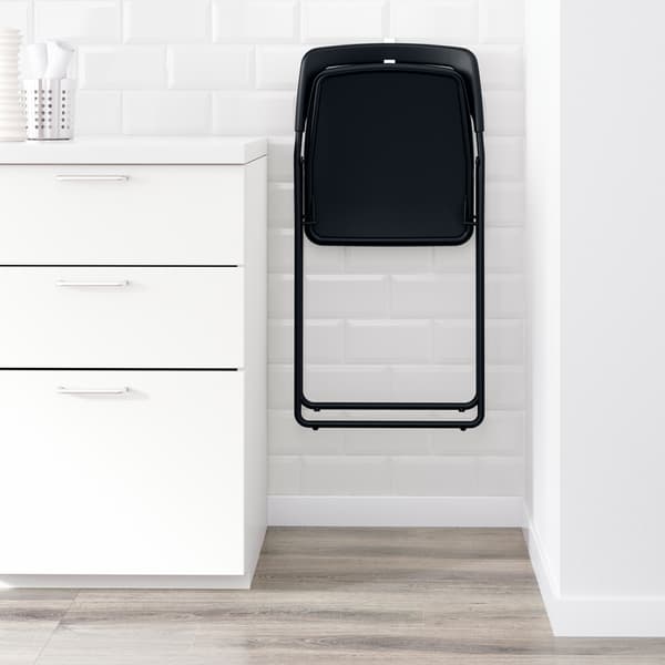 Une chaise pliante noire NISSE suspendue à un mur blanc dans un espace étroit entre un mur et une armoire.