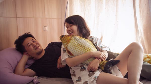 Une chambre à coucher lumineuse où un jeune couple rit et se détend dans un lit avec un oreiller jaune en forme de poisson.