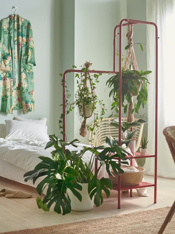 Une chambre avec de petites et grandes plantes vertes suspendues à un portant NIKKEBY rouge placé au bout du lit.
