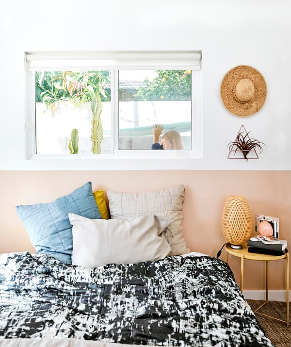 Une chambre avec housse de couette en noir et blanc, des livres et des lampes sur une table de chevet, et Chloé passant devant la fenêtre derrière le lit.