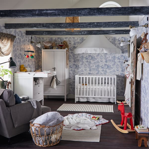 Une chambre d’enfant avec du papier peint bleu/blanc, un lit bébé blanc et une table à langer, une garde-robe blanche et des tapis rayés blanc/noir.