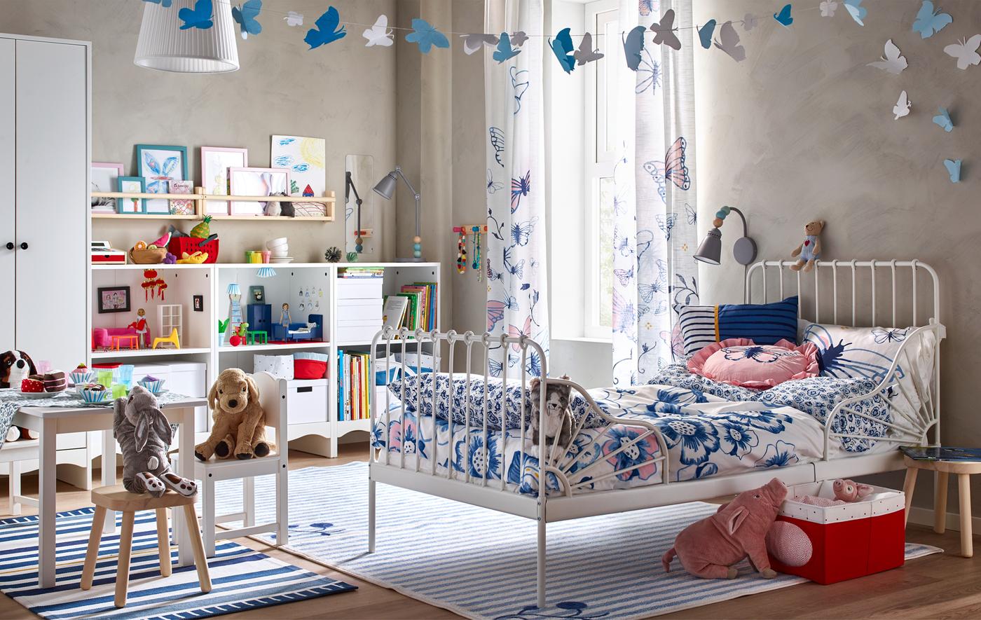 Une chambre d'enfant avec un lit blanc, des textiles floraux, une petite table et des chaises, et un meuble de rangement rempli de livres, de boîtes, et de jouets.