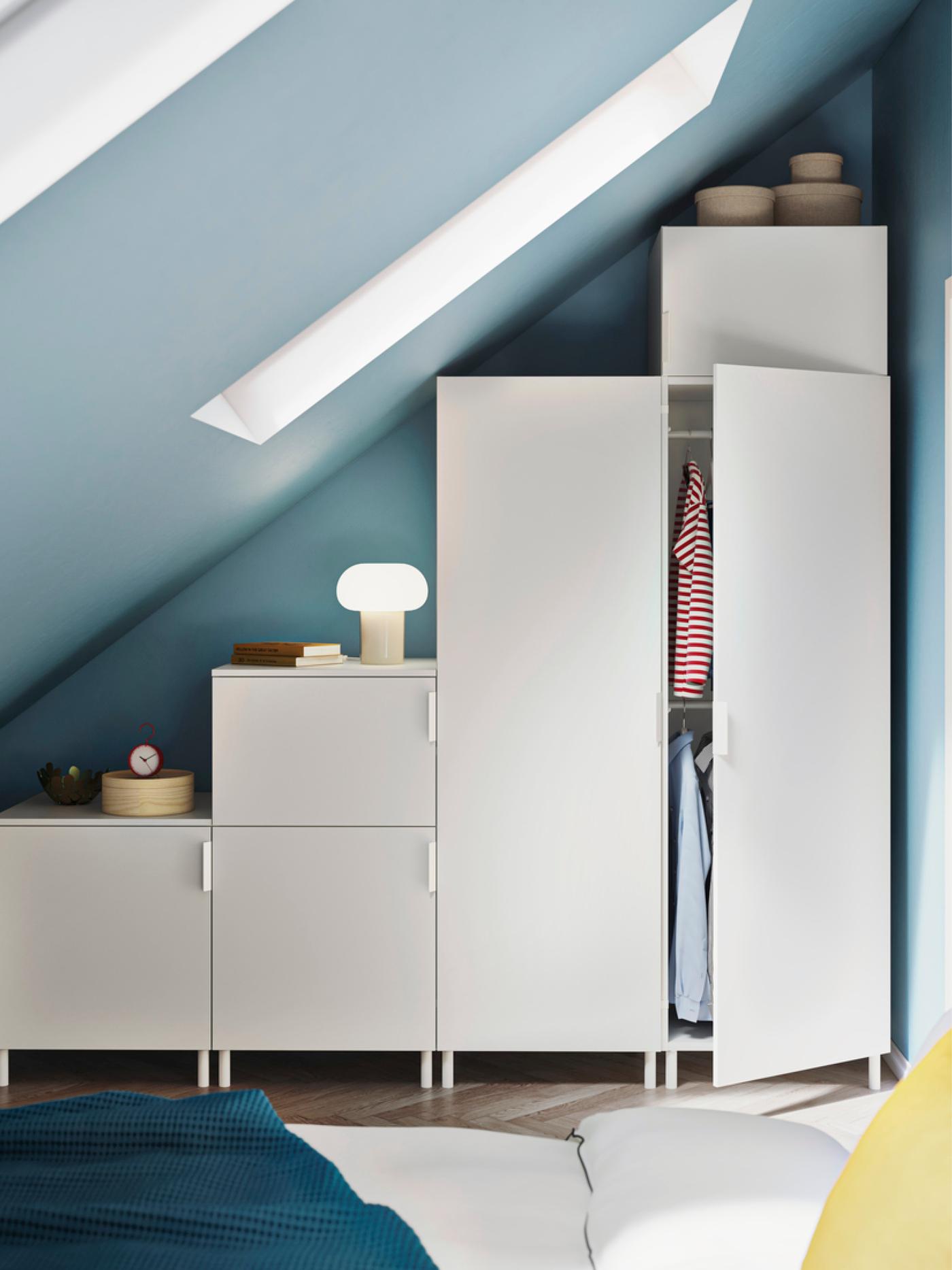 Une chambre moderne avec un plafond en pente et des murs bleus, des armoires modulaires blanches, une armoire ouverte sur laquelle sont suspendus des vêtements et un lit avec du linge de lit blanc, une couverture bleue et un oreiller jaune.