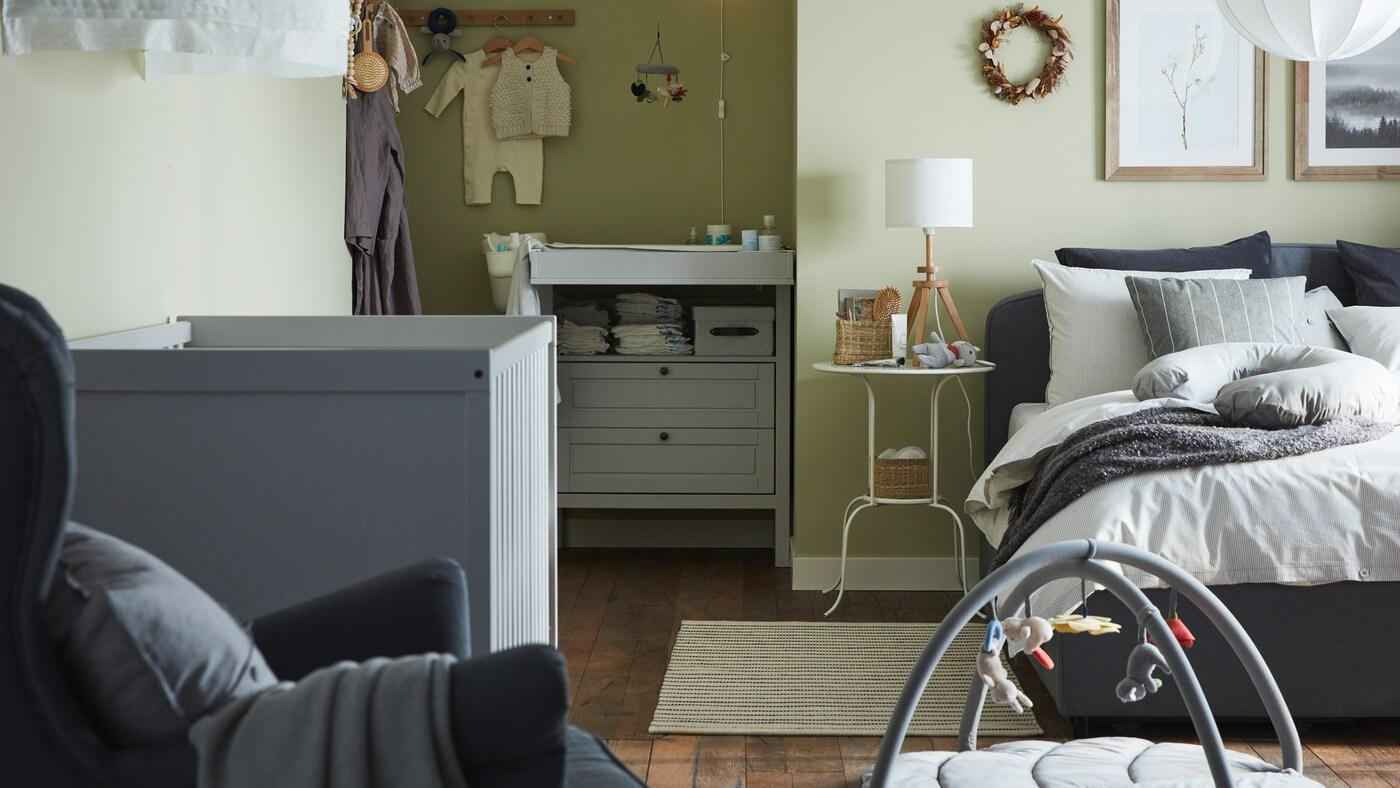 Une chambre pour parents et bébé avec un lit matelassé gris, un lit à barreaux gris, une commode/table à langer grise et des murs verts.