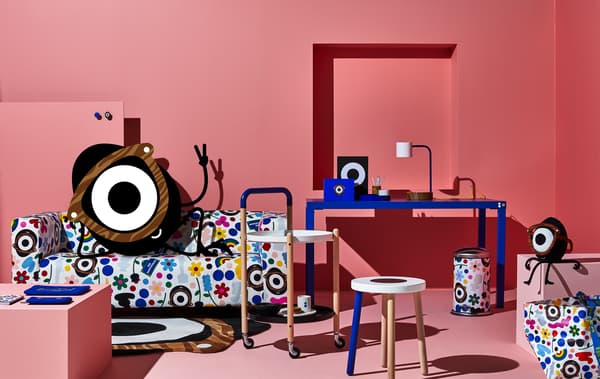 Une chambre rose avec un bureau bleu, une table d'appoint et un tabouret et un cyclope malicieux installé dans un canapé aux motifs multicolores.