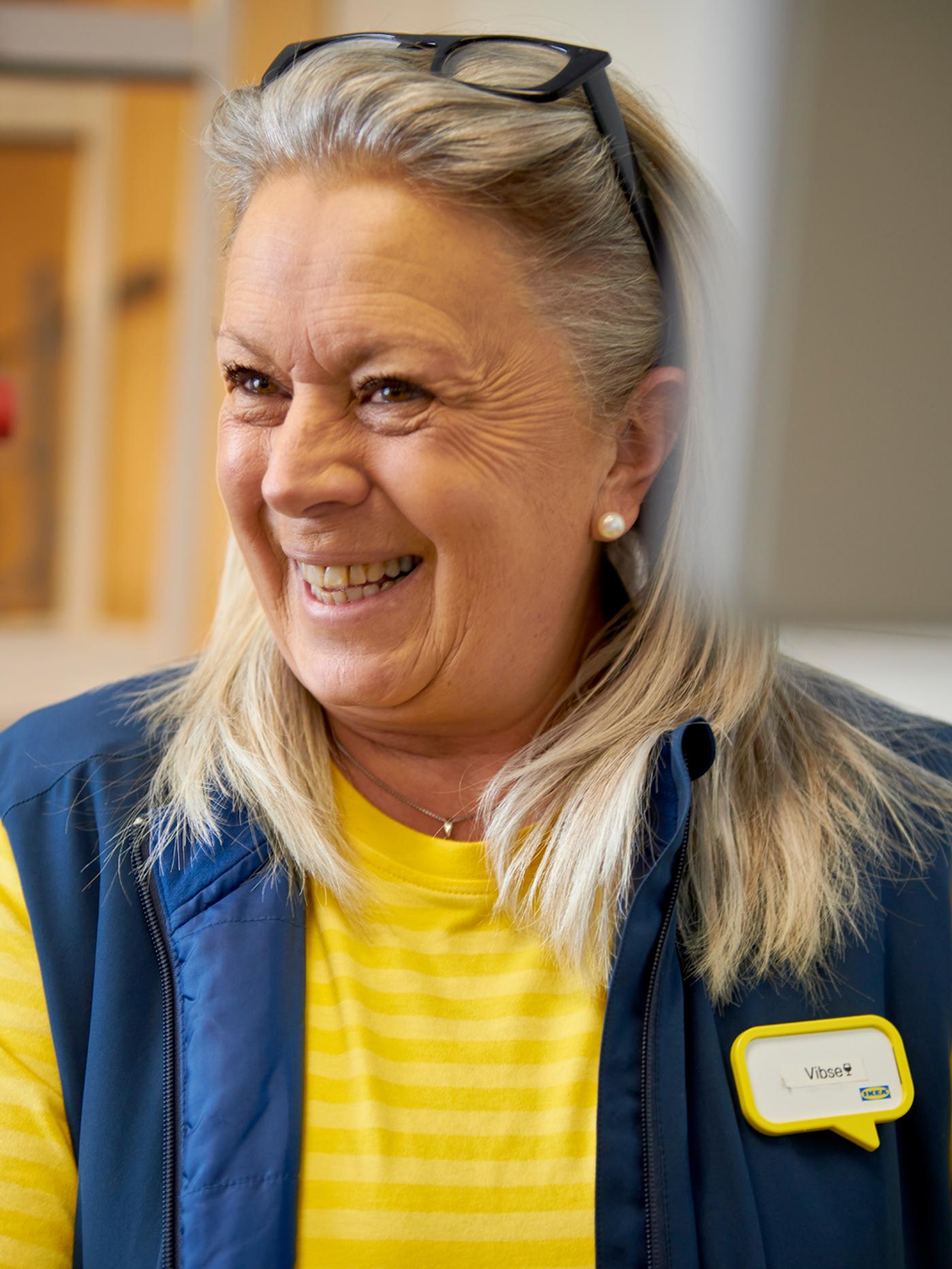 Une collaboratrice IKEA qui porte un chandail jaune et une veste bleue, souriant avec ses lunettes relevées sur la tête.