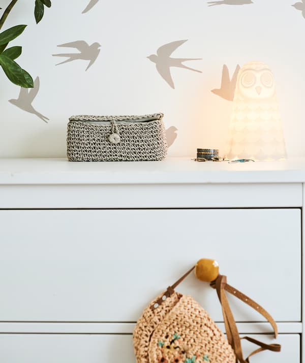 Une commode à tiroirs blanche avec des poignées ambre et une lampe hibou dessus, poussée contre le mur avec des motifs en forme d’oiseaux.