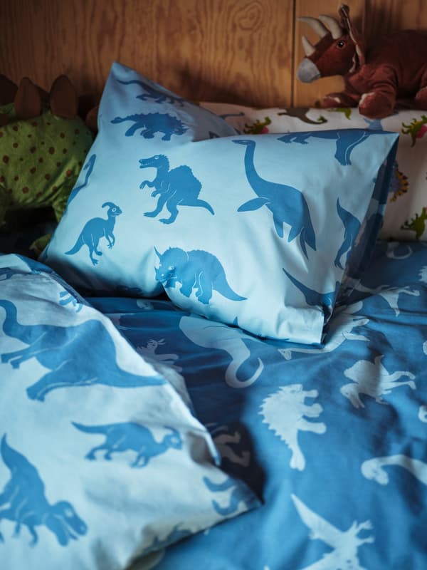 Une couette bleu clair et bleu foncé aux motifs de dinosaures sur un lit d’enfant. Deux dinosaures JÄTTELIK montent la garde.