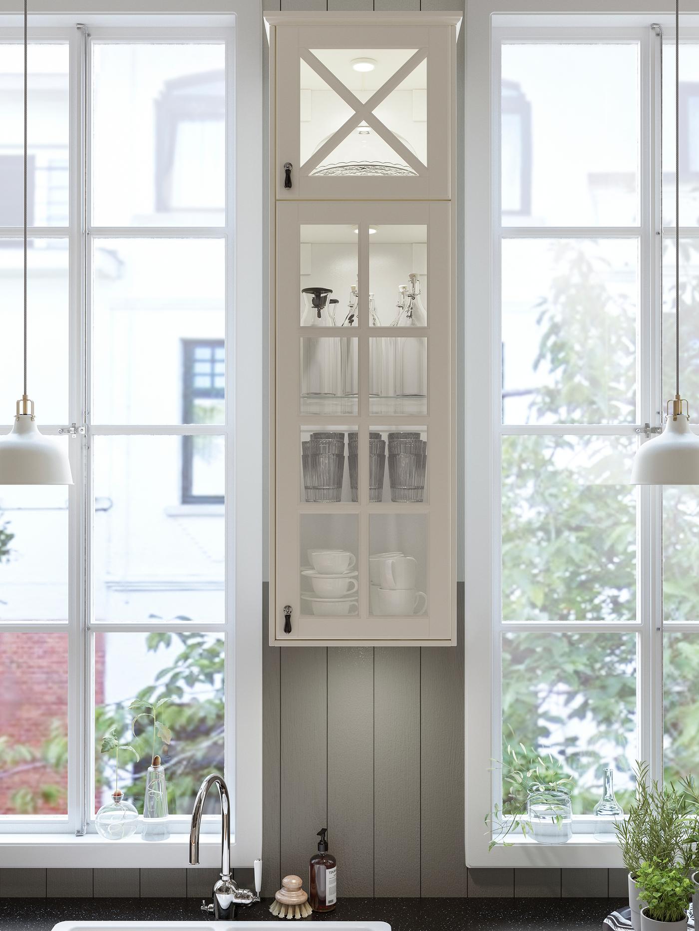 Une cuisine avec une armoire étroite blanc cassé à porte vitrée entre deux fenêtres et un plan de travail à effet minéral noir.