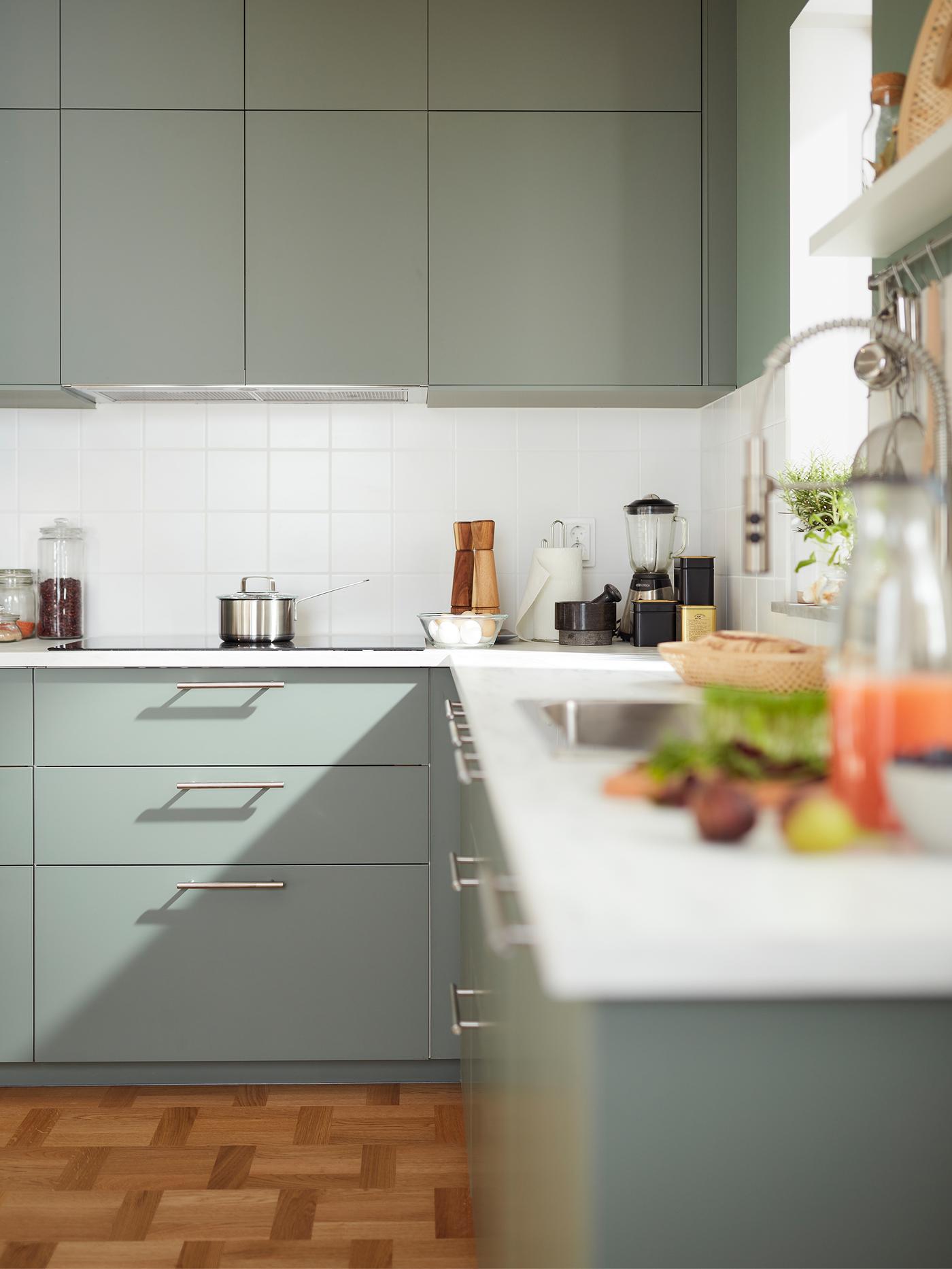 Une cuisine d’angle avec des armoires gris-vert, une plaque de cuisson et un plan de travail à effet minéral gris clair où sont déposés divers ustensiles et appareils.