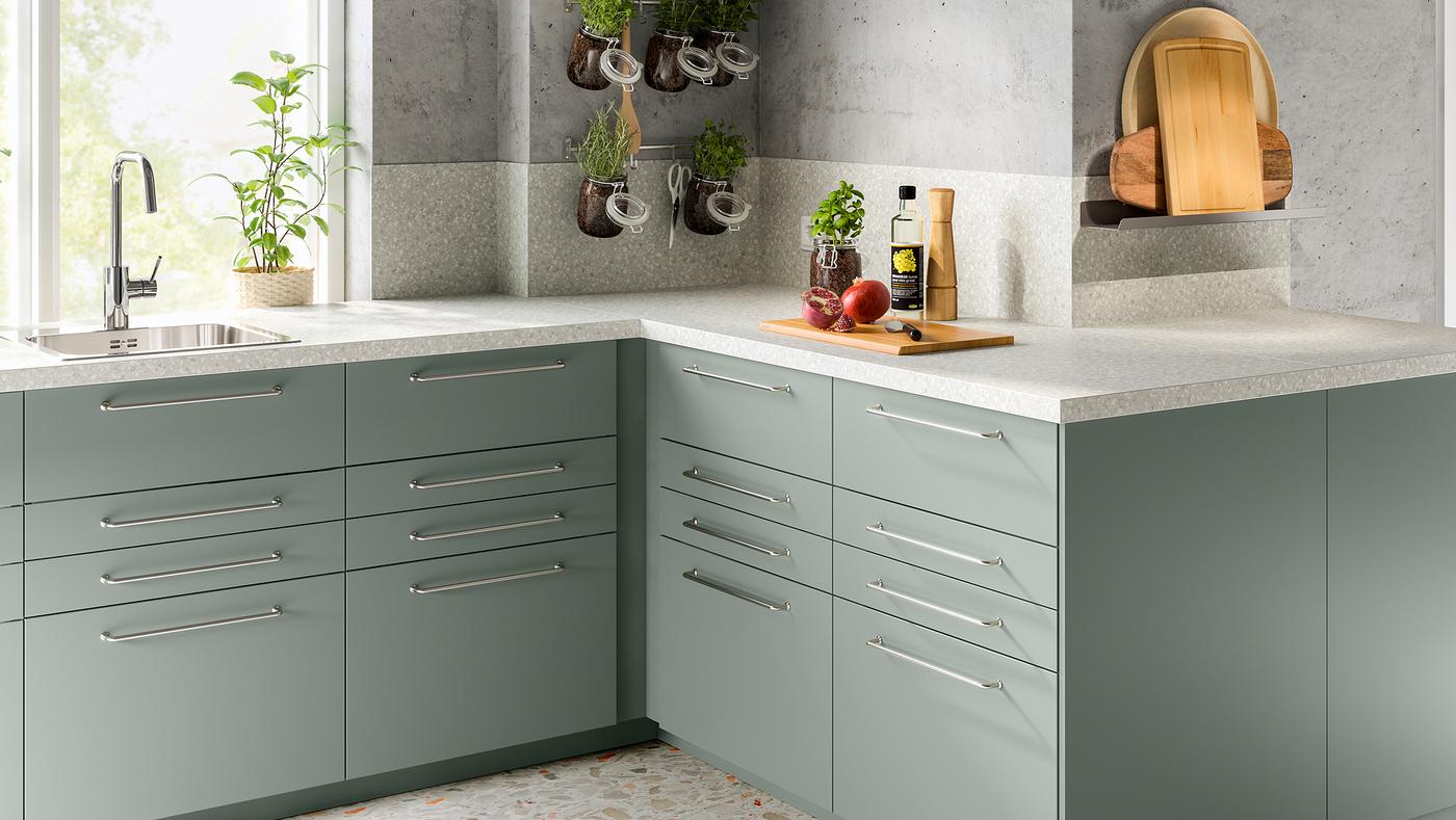 Une cuisine d’angle avec un plan de travail à effet minéral gris clair et plusieurs armoires dont les façades de tiroirs sont revêtues d’un film gris-vert.