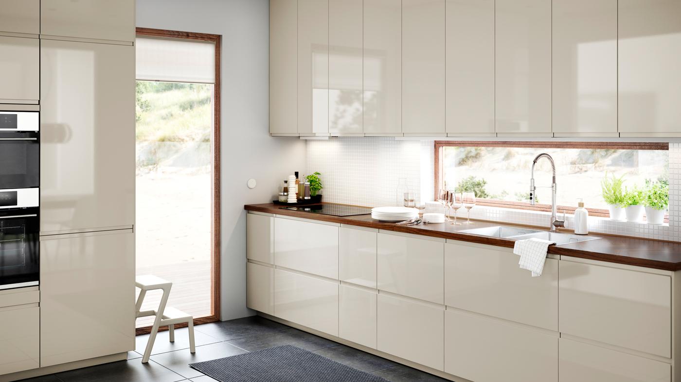 Une cuisine VOXTORP lumineuse et spacieuse, de couleur beige clair, avec des portes et des façades brillantes, et un plan de travail BARKBODA en placage de noyer. portes et façades brillantes, et un plan de travail BARKBODA en placage de noyer.