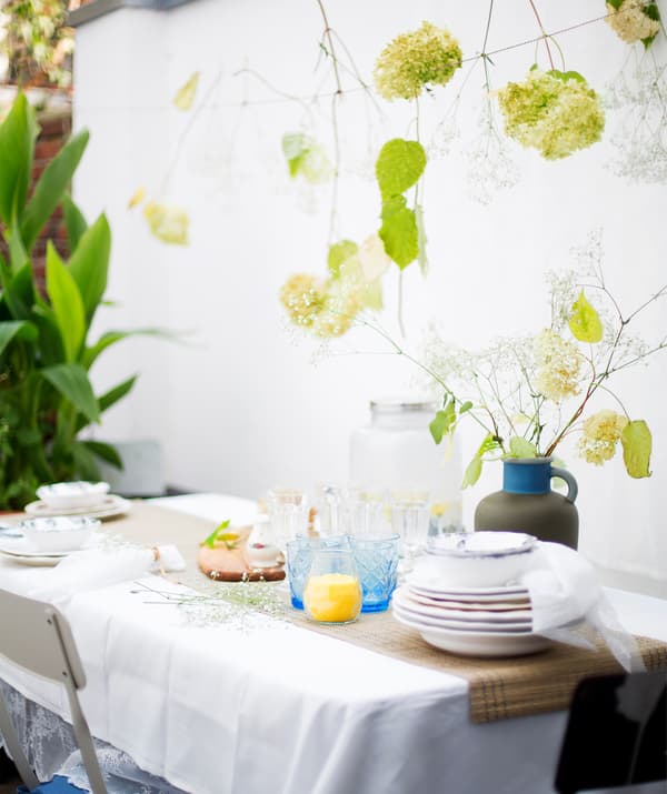 Une décoration de table avec une pile d’assiettes, des verres et un vase de fleurs, avec des fleurs suspendues à de la ficelle fil au-dessus.