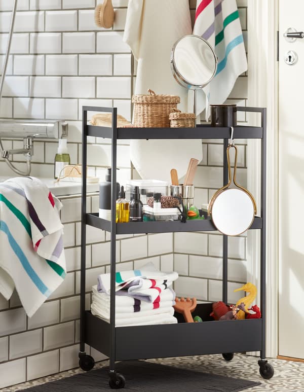 Une desserte en acier NISSAFORS noire avec trois tablettes remplies de jouets de bain, de serviettes, de produits de spa, et des organiseurs tressés en jonc de mer.