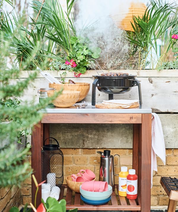 Une desserte en bois dans un coin cuisine à l’extérieur, avec un BBQ sur son plateau en métal et de la vaisselle et des condiments sur une tablette en dessous.