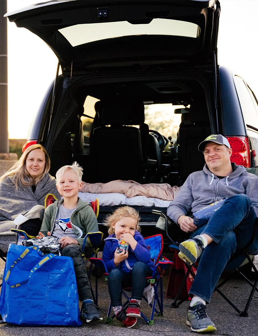 Une famille de quatre personnes est assise sur des chaises de camping derrière leur VUS, prête à regarder le film pendant la soirée ciné-parc.