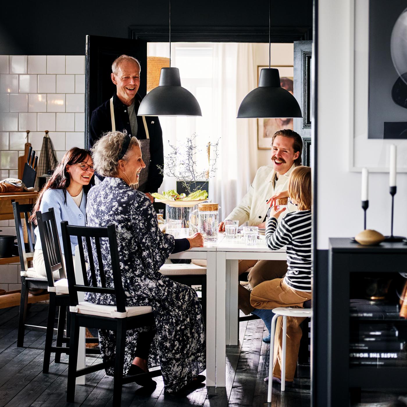 Une famille souriante est assise sur des chaises STEFAN brun-noir autour de deux tables MELLTORP blanches dans une salle à manger en noir et blanc.