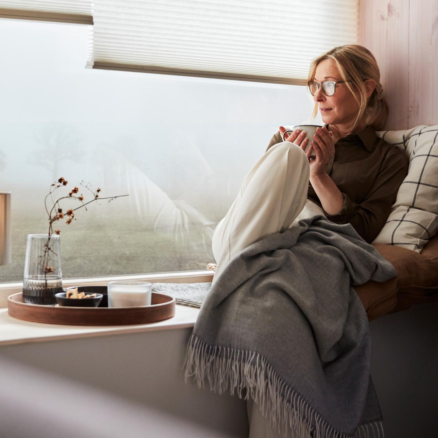 Une femme assise à une fenêtre profitant d'une pause appuyée sur un coussin avec housse SPIKKLUBBA, avec un jeté HOLMVI.