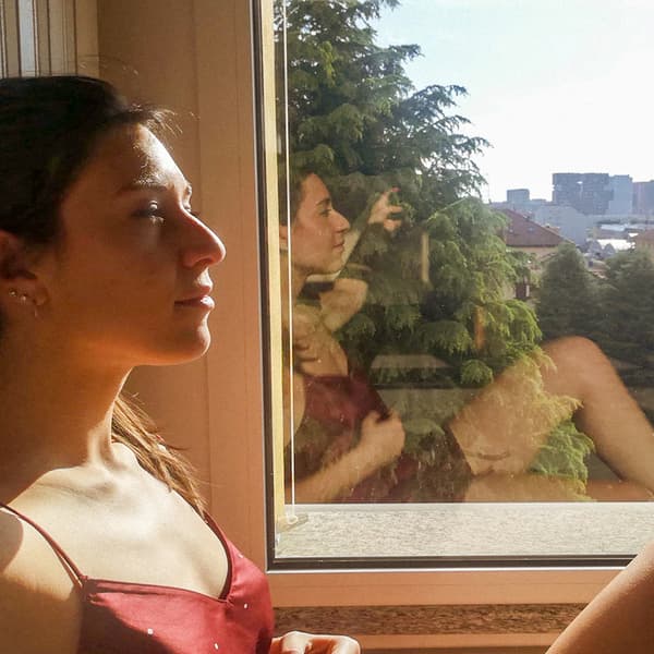 Une femme assise les yeux fermés près d'une fenêtre ensoleillée. Son reflet est visible sur la vitre de la fenêtre derrière elle.