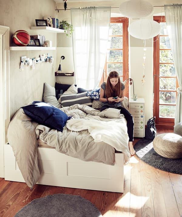 Une femme assise sur un lit double avec des plaids et des coussins dans une chambre à coucher avec de grandes fenêtres.
