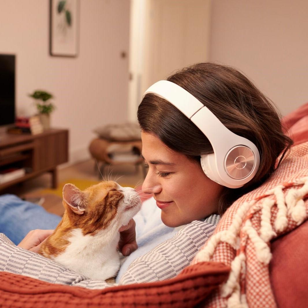 Une femme portant un casque audio est assise sur un canapé et caresse son chat, couché sur son ventre.