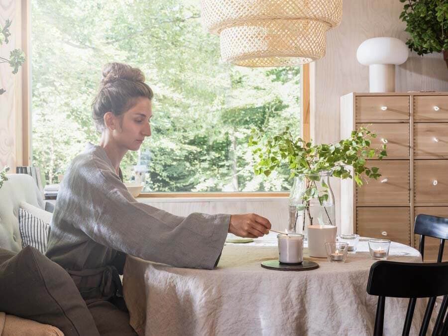Une femme portant un peignoir gris est assise à une table de cuisine à côté de bougies près d’une fenêtre ensoleillée garnie de plantes vertes.