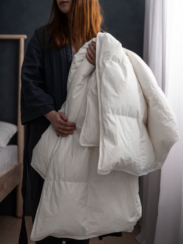 Une femme près d’un lit, dans une chambre faiblement éclairée avec des murs gris agrémentés de voilages blancs, tient une couette FJÄLLARNIKA.
