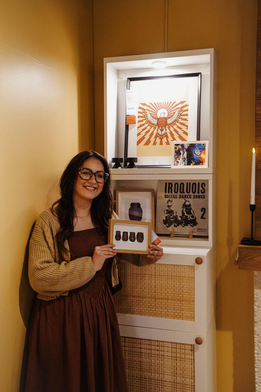 Une femme se tient devant des œuvres d’art avec une œuvre encadrée à la main.