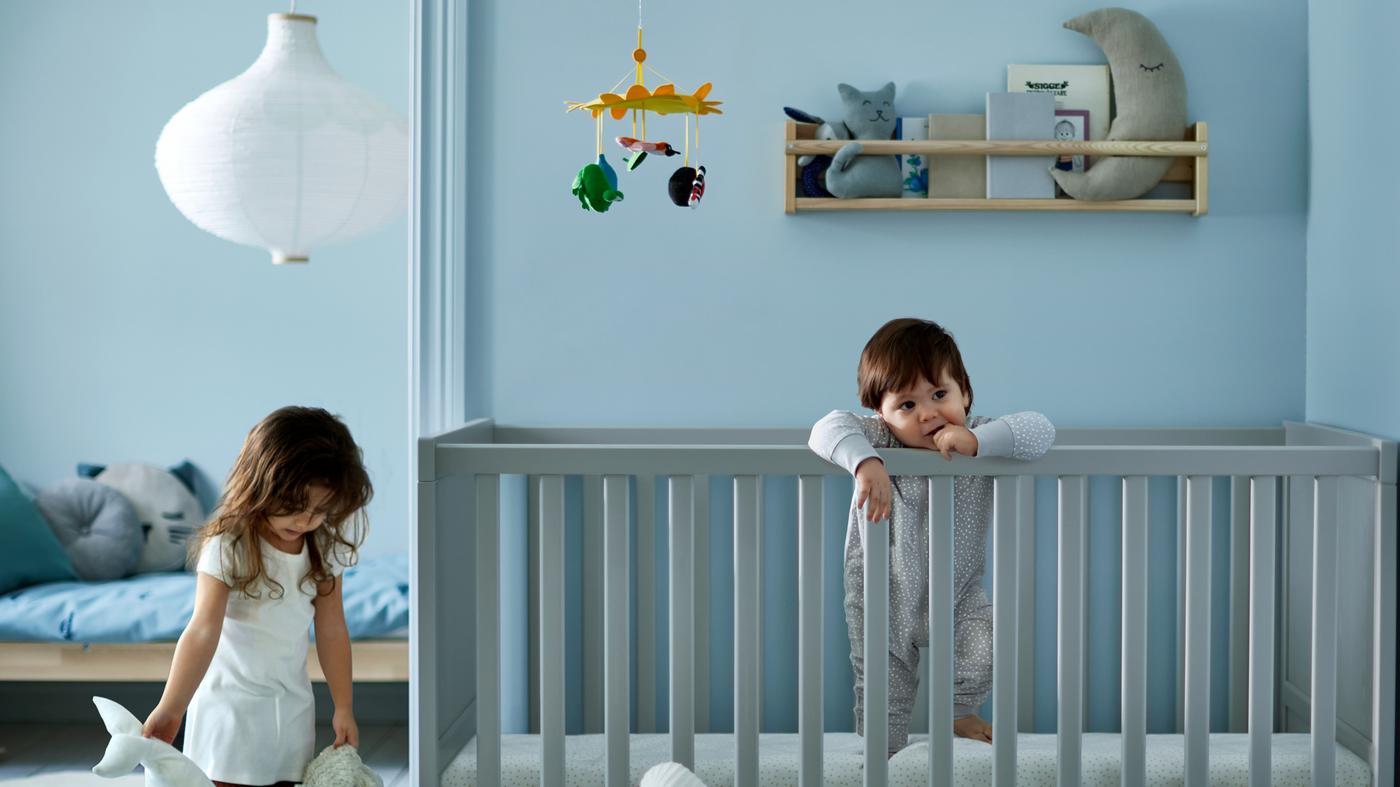 Une fille qui tient deux peluches à côté d'un tout-petit debout dans un lit à barreaux gris, devant un lit pour enfants avec un linge de lit bleu.