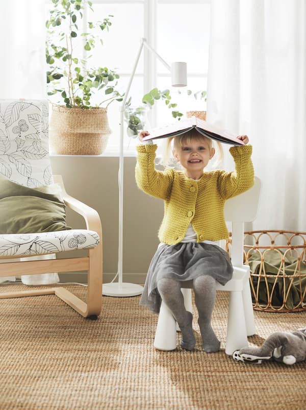 Une fillette utilisant un livre ouvert comme un chapeau est assise sur une chaise pour enfants MAMMUT blanche, placée près d’un fauteuil POÄNG.