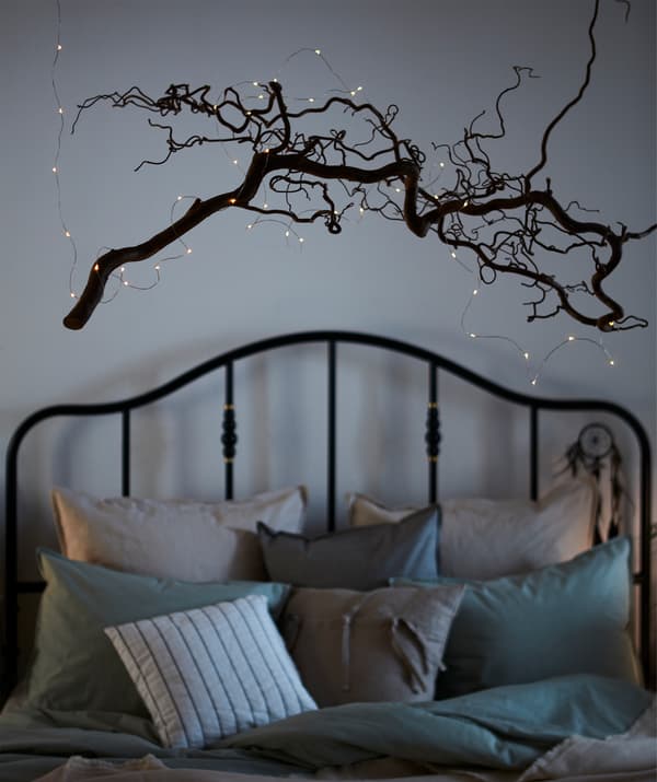 Une fine branche d’arbre torsadée dépourvue de feuilles et parée d’une guirlande VISSVASS accrochée avec du nylon au-dessus d’un lit confortable.