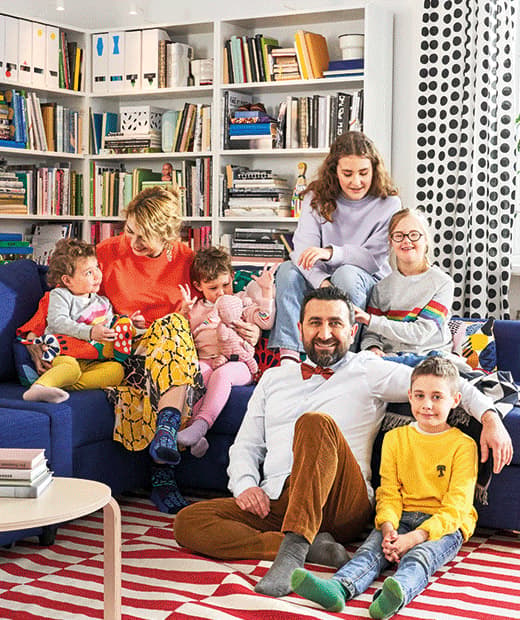 Une grande famille composée des deux parents et de leurs cinq enfants rassemblés autour d’un canapé bleu. Le tapis est rayé rouge et blanc et les bibliothèques en arrière-plan sont bien remplies. 