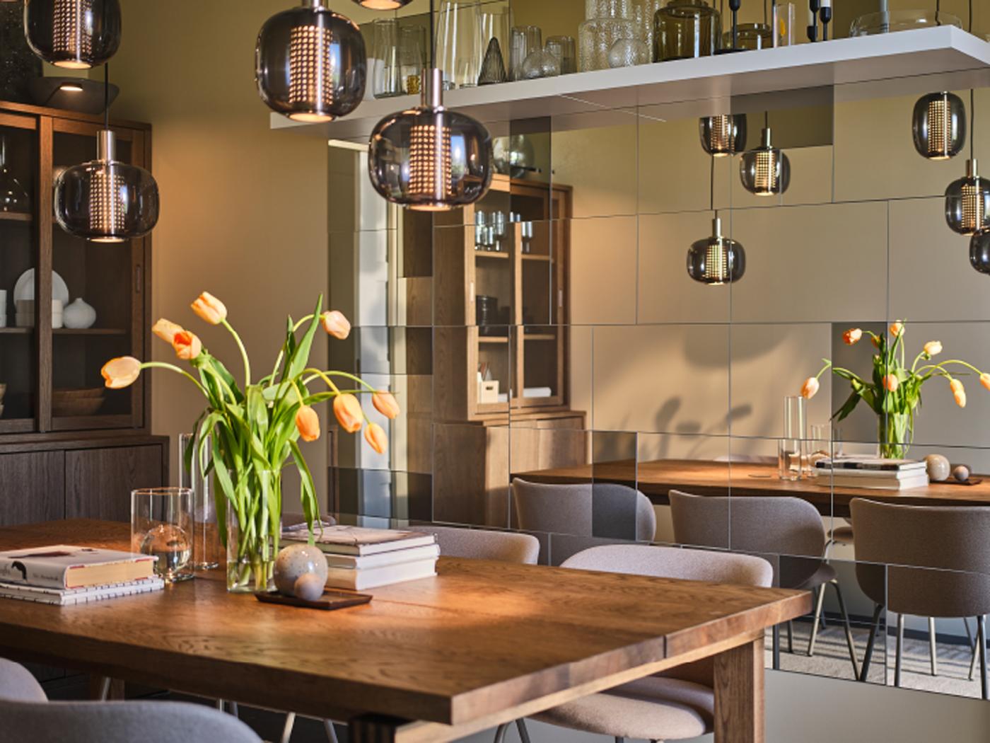 Une grande table MÖRBYLÅNGA sur laquelle sont posés des vases, des fleurs et des livres dans une salle à manger avec plein de miroirs sur un mur.