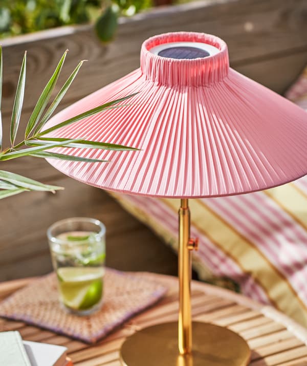 Une lampe de table à énergie solaire SOLVINDEN, éclairée par le soleil, posée sur une petite table, à côté d’un verre contenant une boisson et des rondelles de citron. 