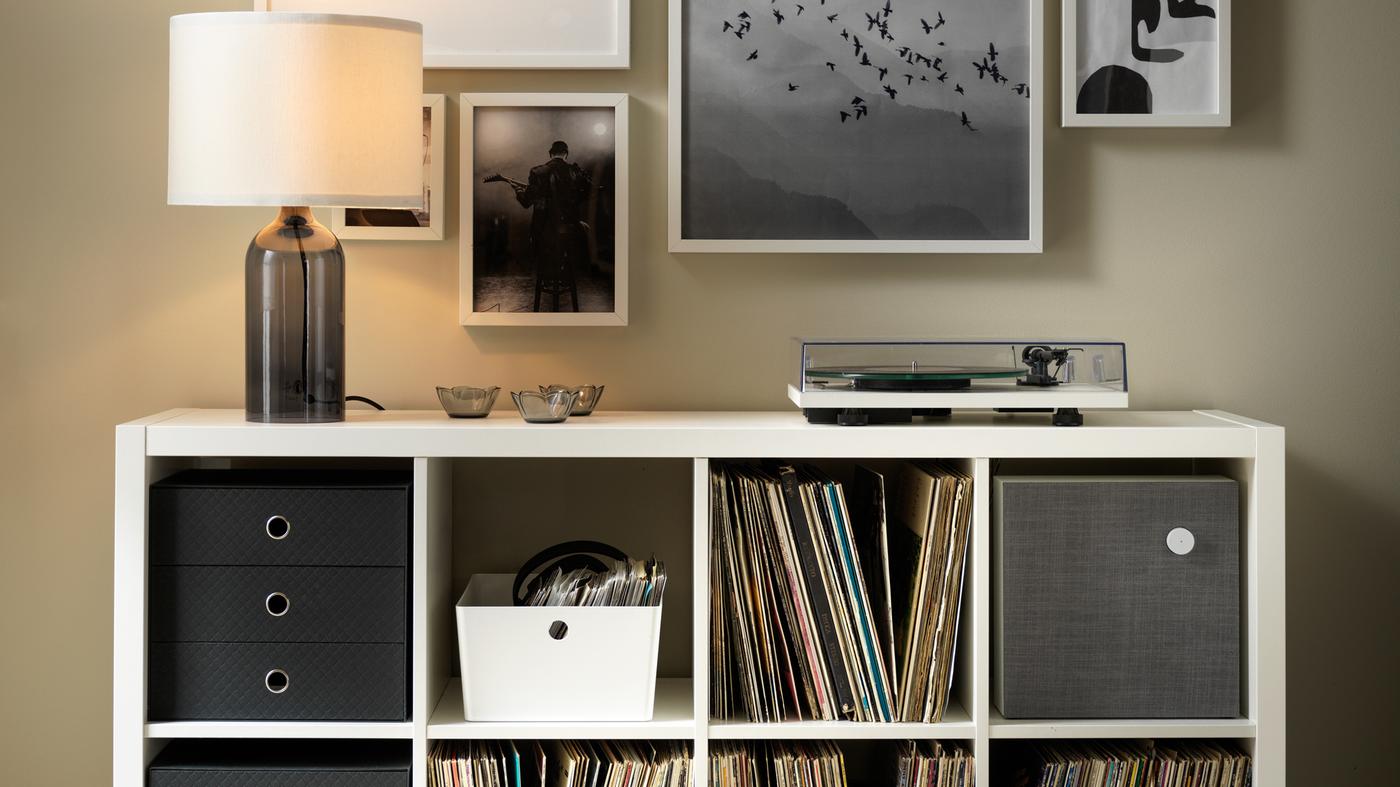 Une lampe de table TONVIS et un tourne-disque sur une étagère KALLAX blanche remplie de disques vinyles et divers autres petits rangements.