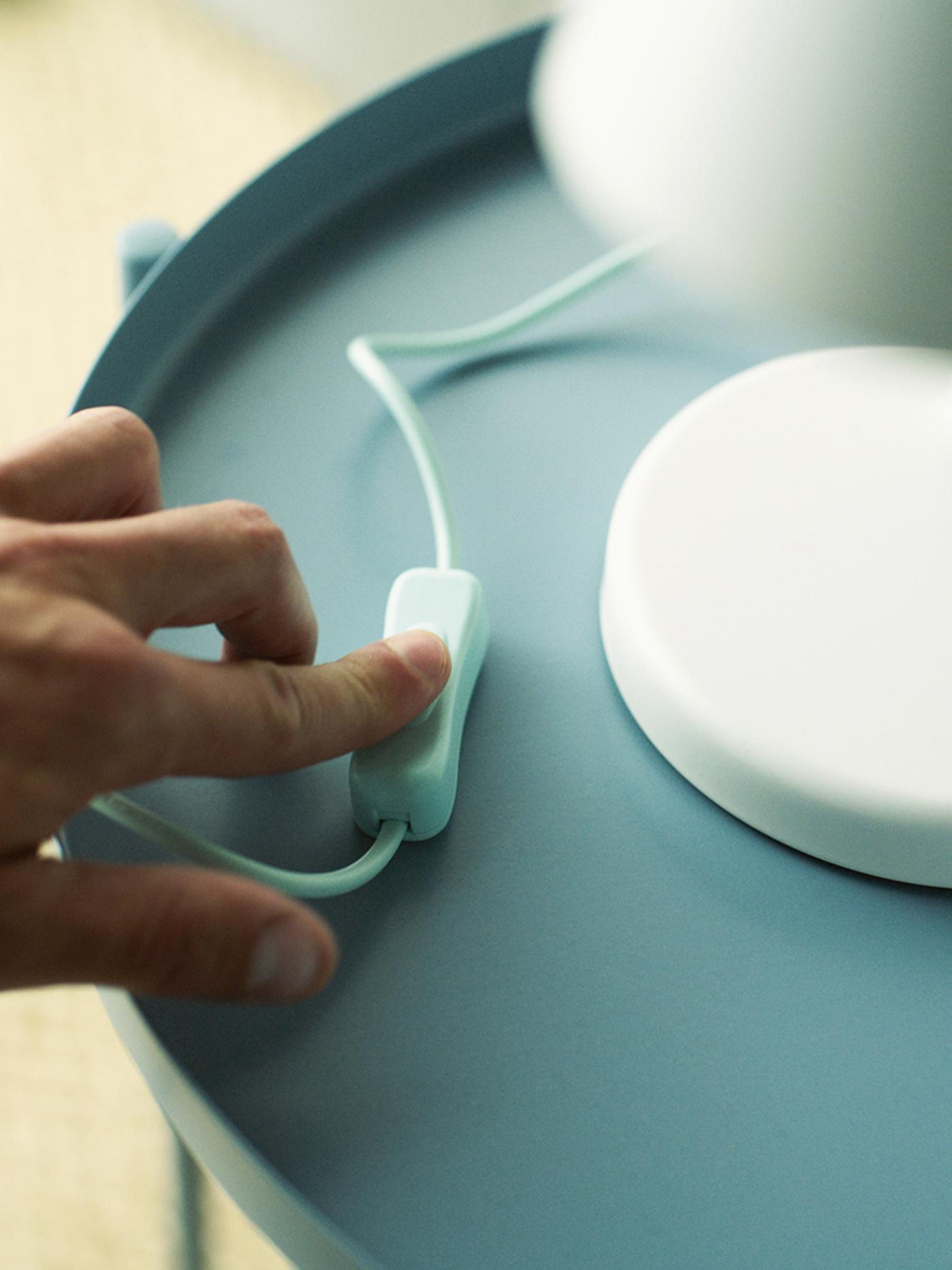 Une main actionne l’interrupteur d’une lampe de table blanche posée sur une table bleu clair.